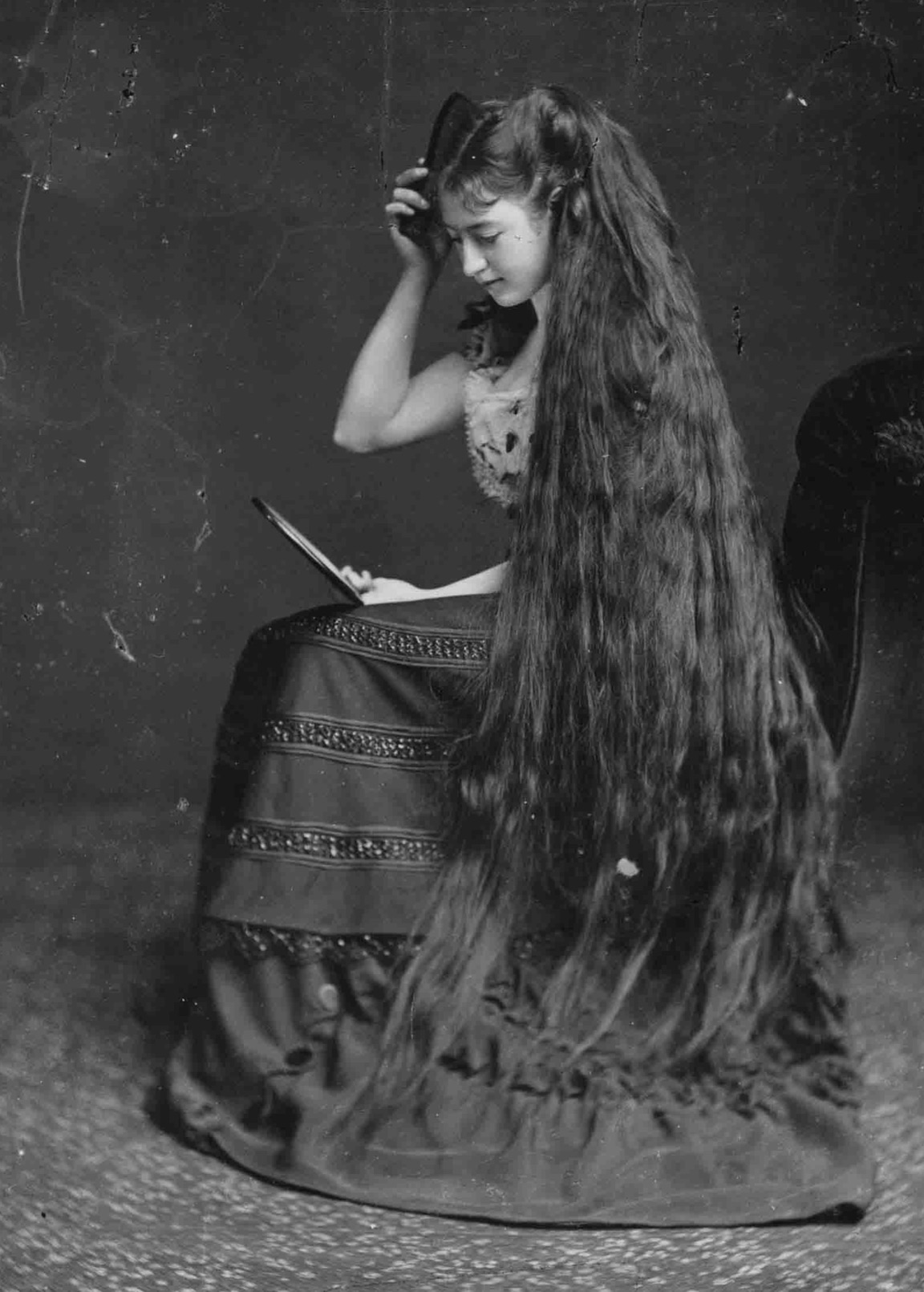 Woman with long hair, brushes it off while looking at the mirror, 1880s